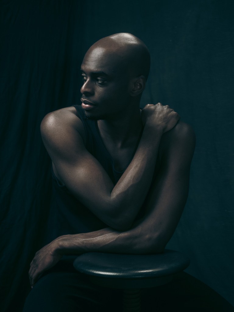 Fine art portrait of Kioff by De Sheng Lim, a portrait photographer based in The Hague, Netherlands. Kioff is seen in a side profile, resting his head near his crossed arms on a stool, showcasing the interplay of light and shadow on his skin.