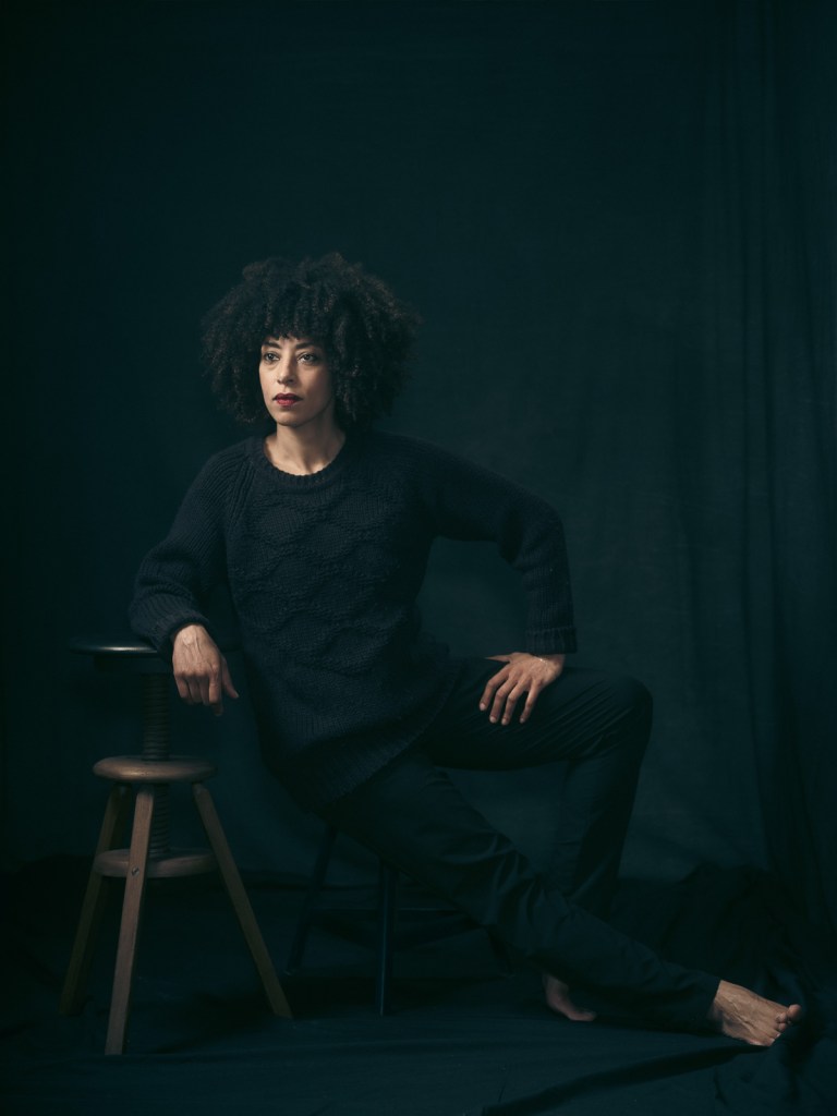 A full-length fine art portrait of Lea leaning against a tall wooden stool in a dark studio setting, captured by De Sheng Lim, a portrait photographer based in The Hague, The Netherlands.